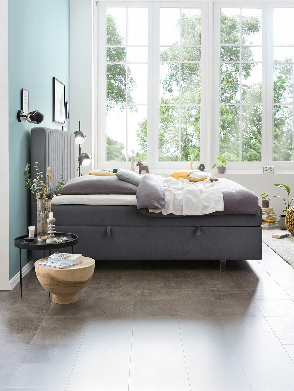 Ein Schlafzimmer mit einem grauen Boxspringbett, einem Holztisch und einem Fenster mit Blick auf Bäume.