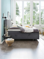 Ein Schlafzimmer mit einem grauen Boxspringbett, einem Holztisch und einem Fenster mit Blick auf Bäume.