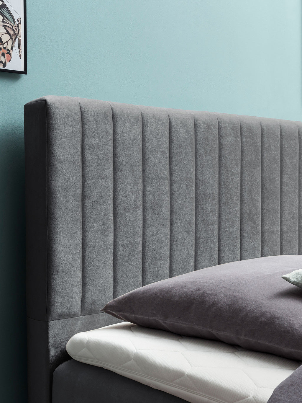 Graues gepolstertes Kopfteil mit vertikalen Nähten, davor ein Kissen auf einer Matratze. Eine blaue Wand im Hintergrund.