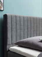 Graues gepolstertes Kopfteil mit vertikalen Nähten, davor ein Kissen auf einer Matratze. Eine blaue Wand im Hintergrund.