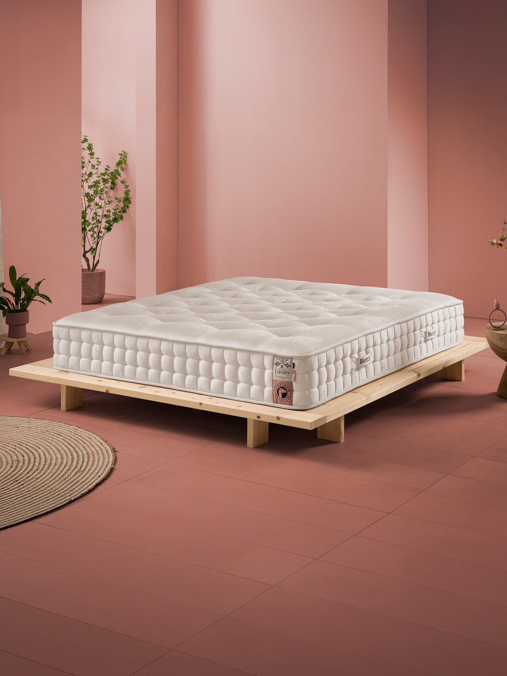 Weiß-beige Matratze auf einem niedrigen Holzrahmenbett in einem Raum mit rosa Wänden und Terrakotta-Boden.