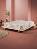 Weiß-beige Matratze auf einem niedrigen Holzrahmenbett in einem Raum mit rosa Wänden und Terrakotta-Boden.